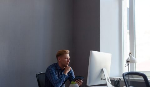 Young casual male sitting in front of computer with tablet in hands in office.
